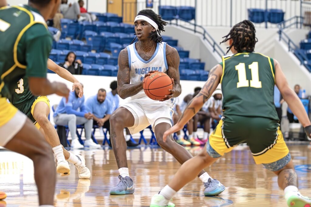 Basketball player in white holds the ball, guarded by two in green on indoor court; spectators and teammates in background.