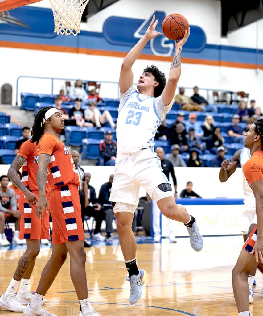 Player in white jersey 23 jumps for a layup as orange team watches on indoor court with spectators in the background.