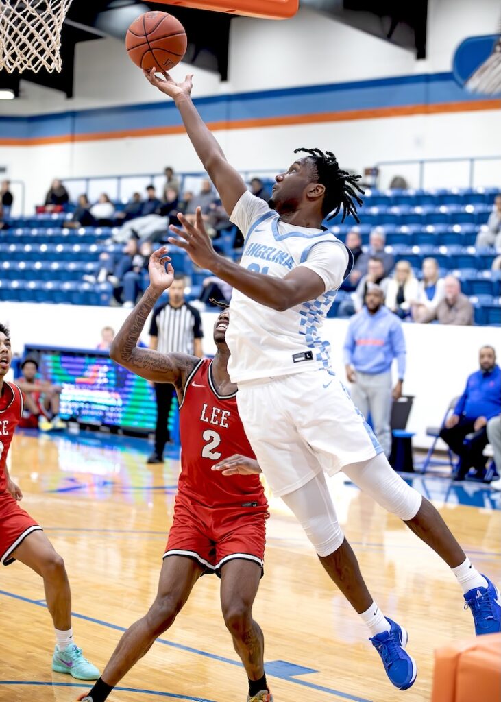 Basketball player in white and blue leaps for a layup as a red-uniformed defender tries to block, gym crowd in background.