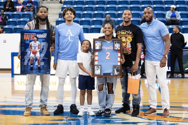 Kai’Ree Murray poses at center court on Sophomore Day with family, holding a framed jersey and player poster.