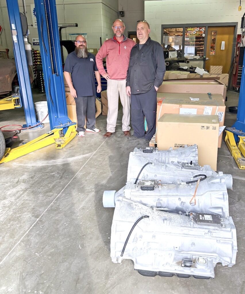 Three men smiling in an auto repair shop with large parts, boxes, and vehicle lifts in the background.