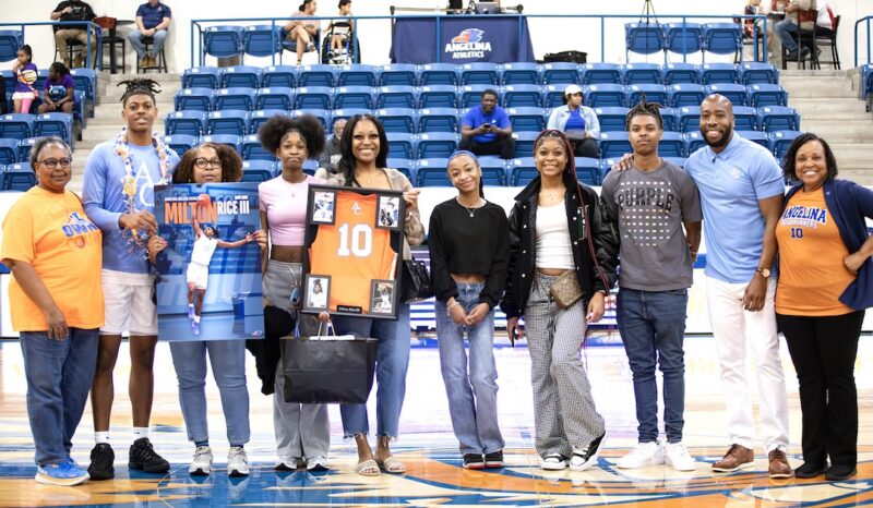 Milton Rice III stands at center court on Sophomore Day holding a framed jersey and poster with family and coaches.