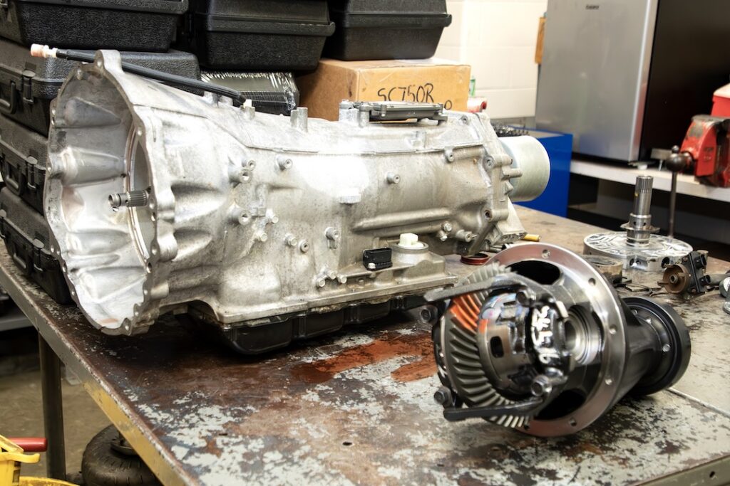 Car transmission and differential gears sit on a workshop bench, surrounded by tools, storage cases, boxes, and parts.