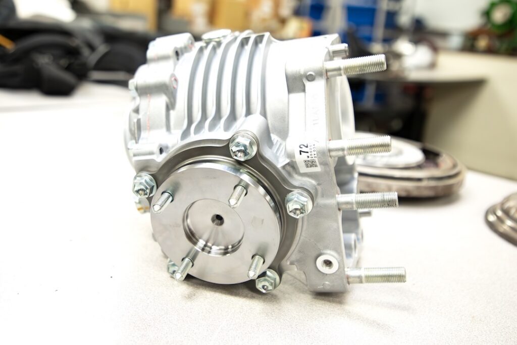 Metallic engine part with bolts and a round silver cover on a light table; blurred tools and machinery in the background.