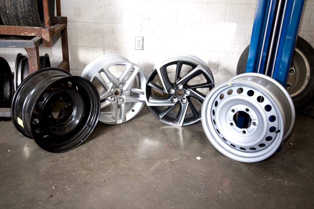Four car rims—two silver with spokes, one black, and one simple silver—are lined up on a concrete floor against a white wall.