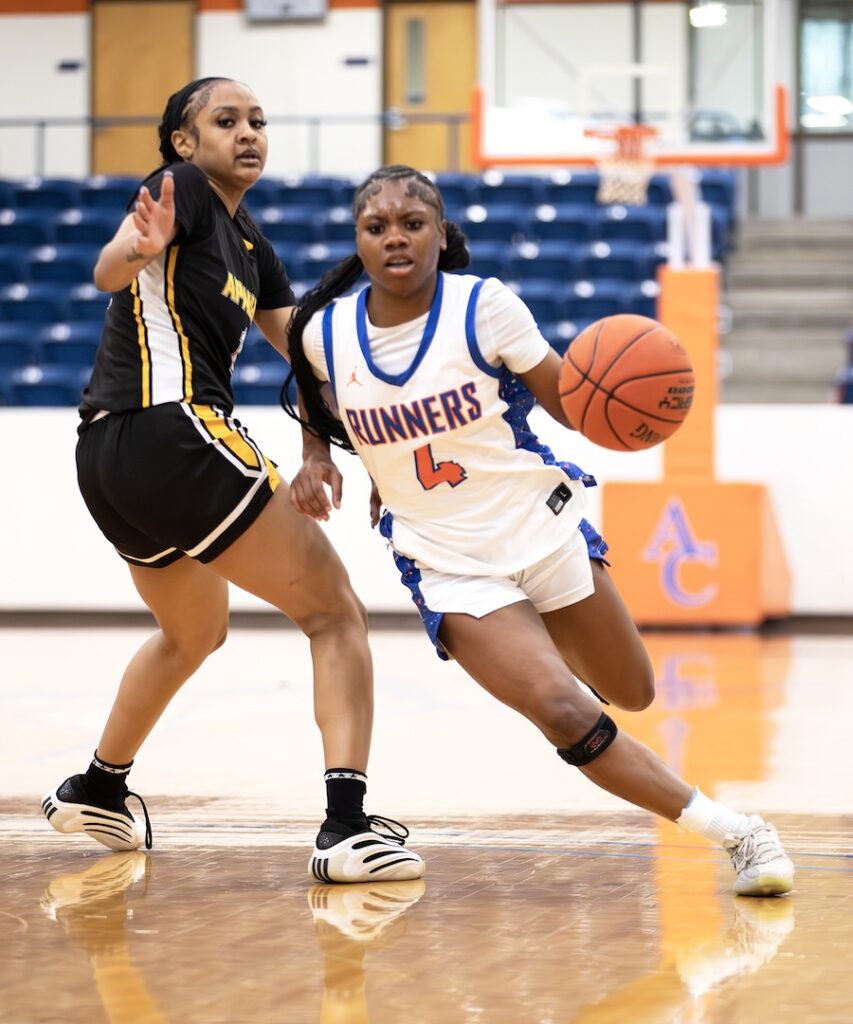 Basketball player in white and blue Runners uniform dribbles past defender in black and yellow on indoor court toward the hoop.