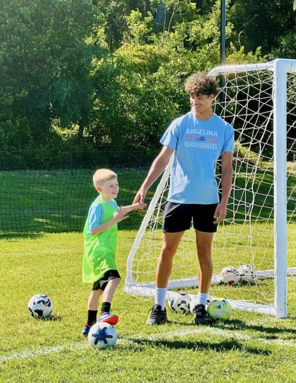 Young boy in green jersey smiles, holding hands with coach in blue by a soccer goal, grassy field, and several soccer balls.