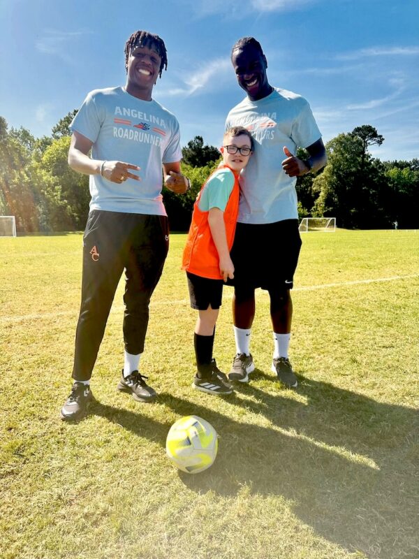 Two adults in Angelina Roadrunners shirts and a smiling child in a red jersey stand on grass by a soccer ball.