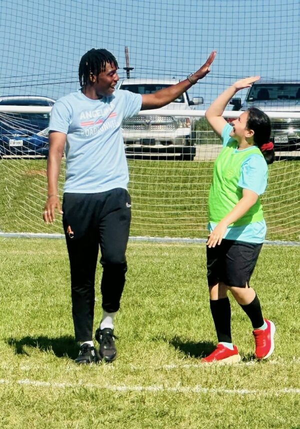 Two people on a soccer field high five and smile in front of the goal, one in blue, the other in a green vest.