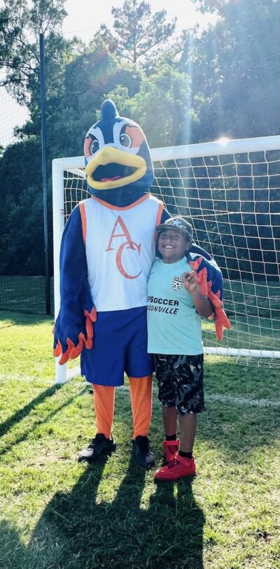 Smiling child makes a peace sign next to a colorful bird mascot in front of a soccer goal on a sunny grassy field.