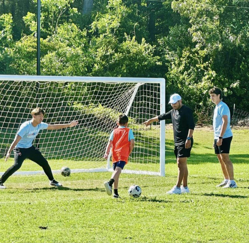 Child in red vest kicks soccer ball at goal as a goalkeeper prepares to block; three adults watch and instruct on sunny field.