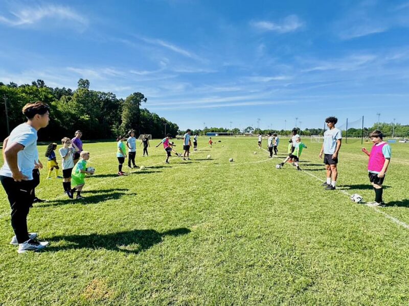 Children and adults line up on a grassy soccer field under a blue sky, training with cones and balls scattered around.