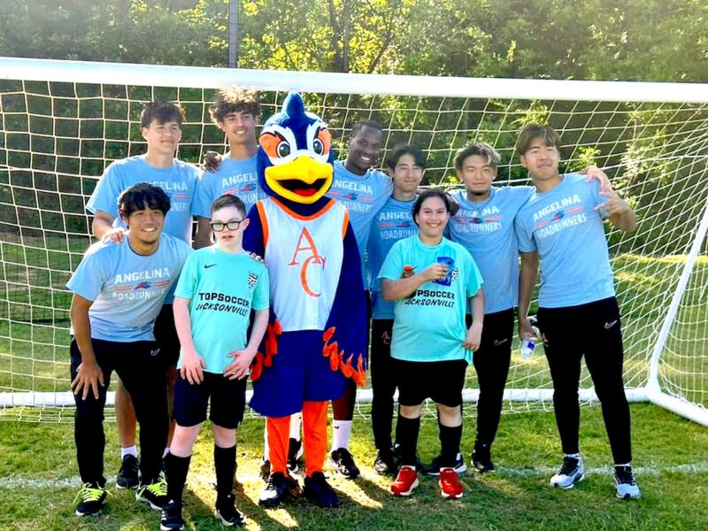 Young soccer players and a bird mascot, all in blue shirts, smile together in front of a goal on a sunny day.