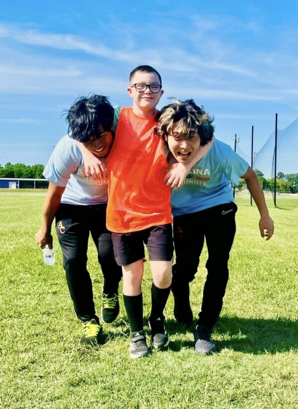 Three boys smile on a grassy field; the center boy in orange hugs two teammates in light blue shirts on a sunny day.