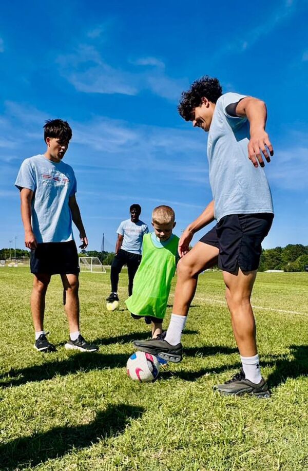 Four boys play soccer on a sunny field; three in blue shirts and black shorts, one younger boy in green vest about to kick the ball.
