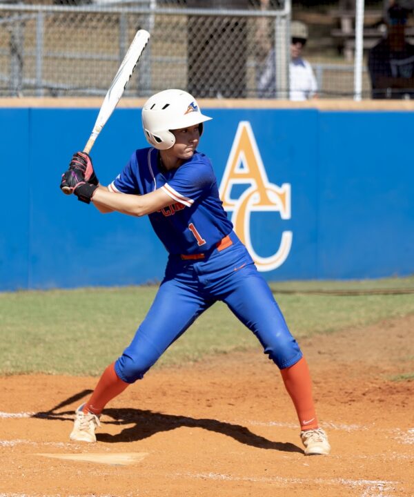Softball player in blue and orange uniform and white helmet prepares to bat on dirt field, blue fence with AC letters behind.