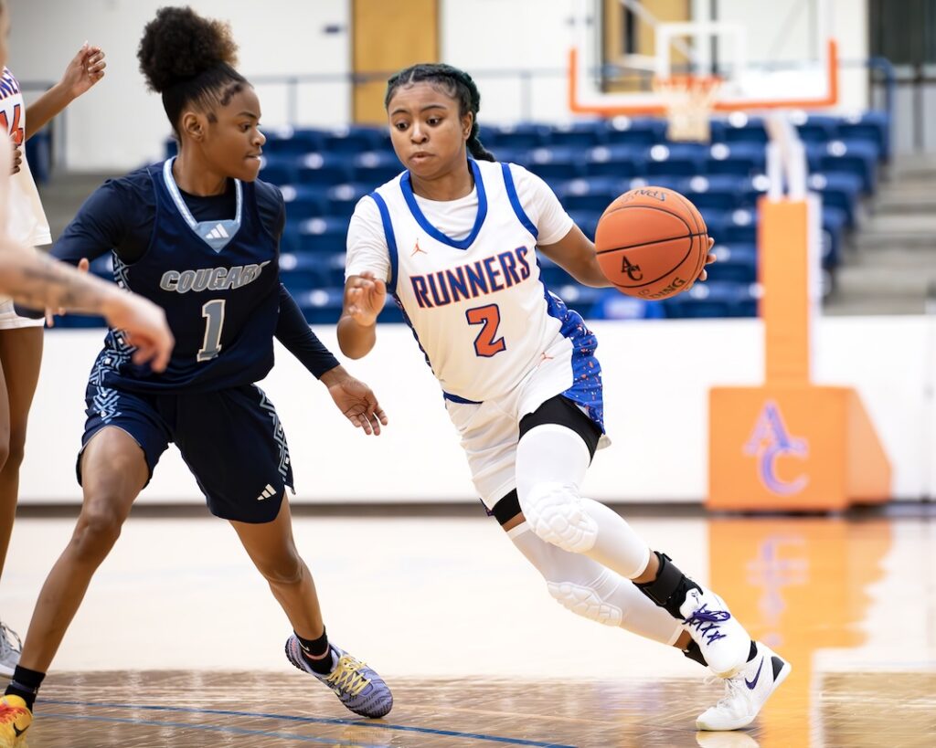 A Runners player in white dribbles while guarded by a Cougars defender in dark; court and empty seats in background.