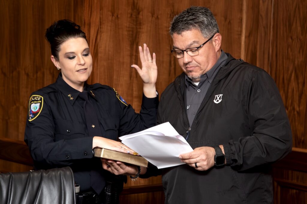 A police officer holds a book as a man, raising his right hand, reads papers and takes an oath in a wood-paneled room.