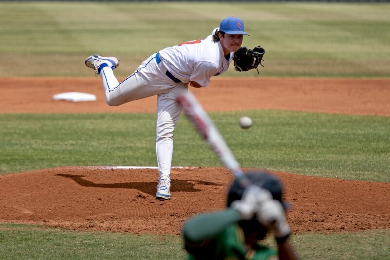 Baseball pitcher in white throws as batter in dark uniform swings at a mid-air ball on a sunny outdoor field.
