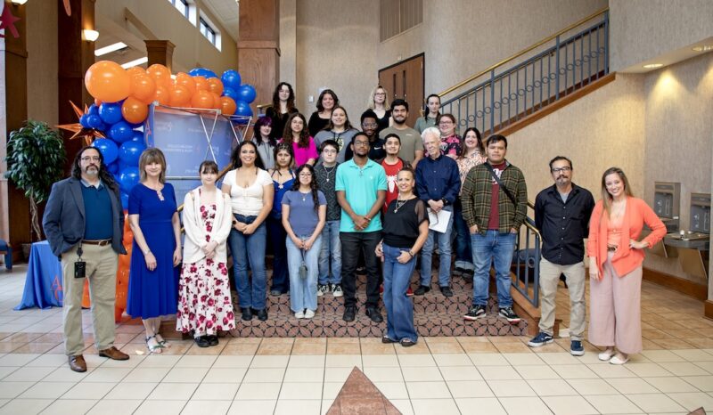 Around 25 people pose on indoor steps, smiling before blue and orange balloons at a formal event in a spacious building.