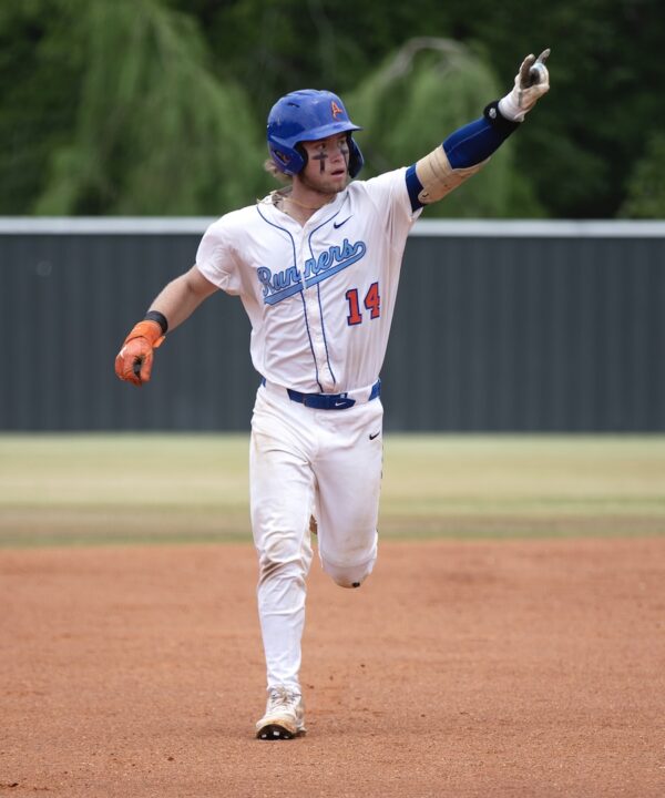 Baseball player in white uniform with blue helmet runs on dirt field, pointing up; trees and a fence in the background.