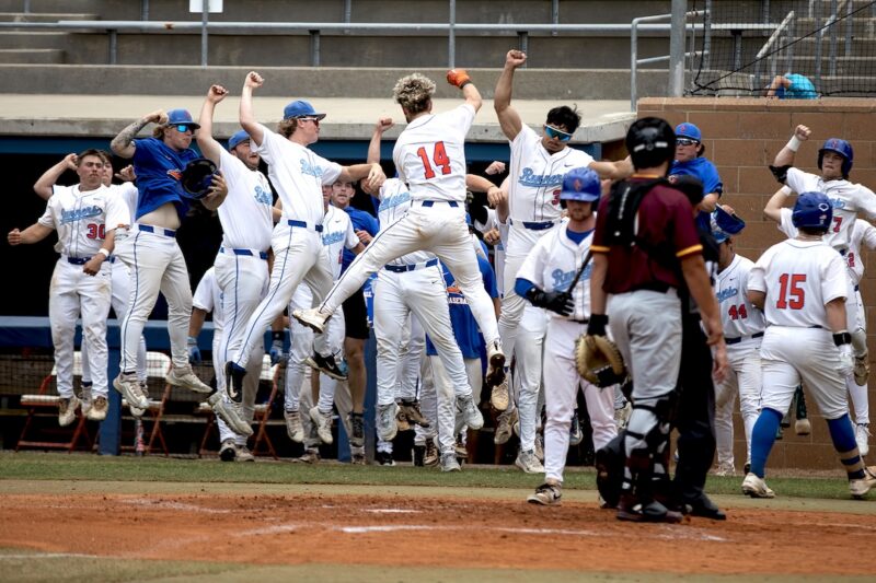 Baseball team in white with blue accents cheers by the dugout as opposing players in maroon and gray stand on the field nearby.