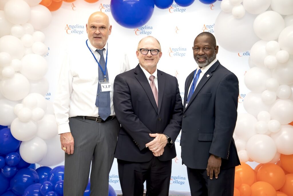 Three men in suits smile in front of an Angelina College logo backdrop with white, blue, and orange balloons; one wears a VIP badge.