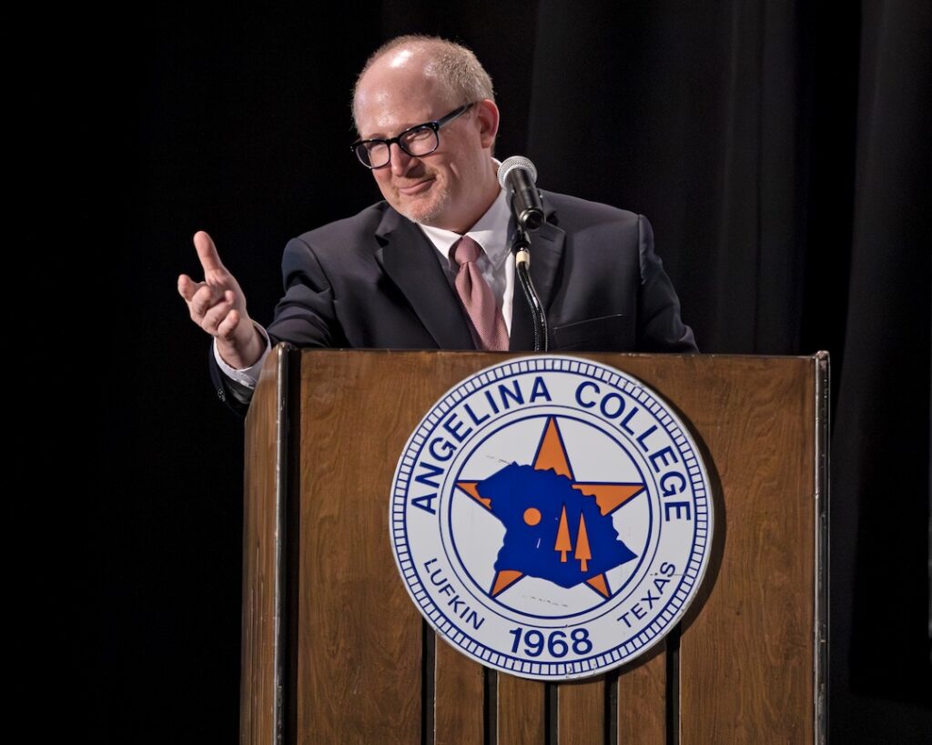 Man in a suit smiles and gestures while speaking at a podium with the Angelina College logo and a microphone.
