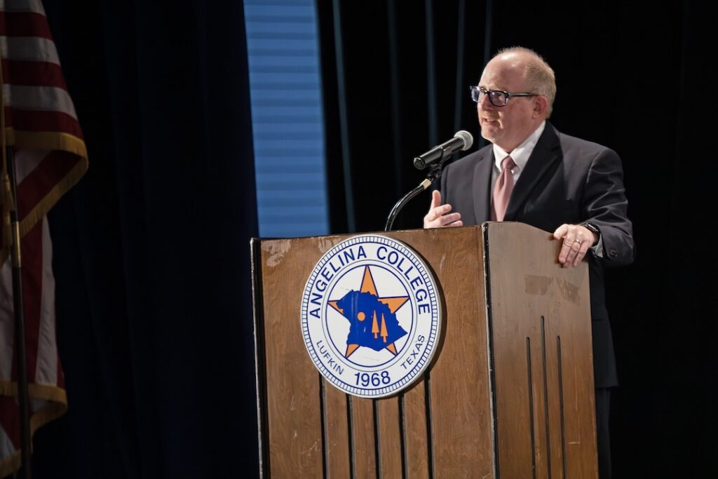 Man in suit and tie speaks at podium with Angelina College seal, microphone, and American flag on stage indoors.