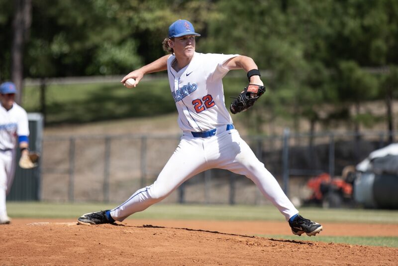 Baseball player in a white uniform, number 22, pitches on a mound; trees and another player are visible in the background.