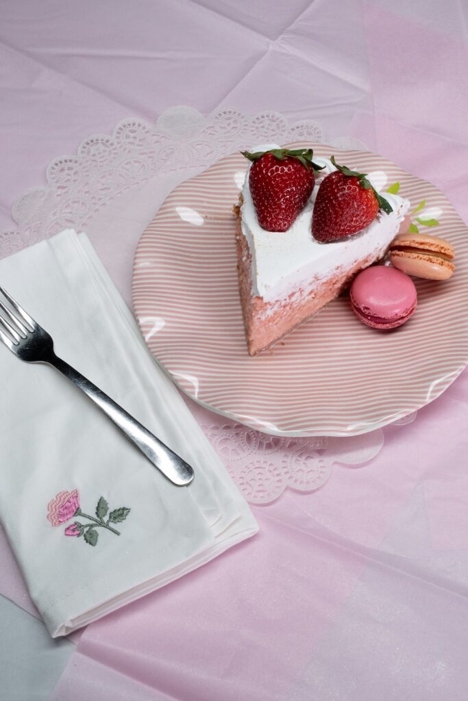 Strawberry cake slice with two strawberries, pink and orange macarons on striped plate, napkin, fork on pink tablecloth.