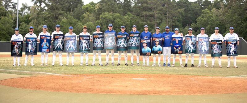 Baseball players in uniform stand in a line on a field, each holding a poster with their photo; forest and trees in background.