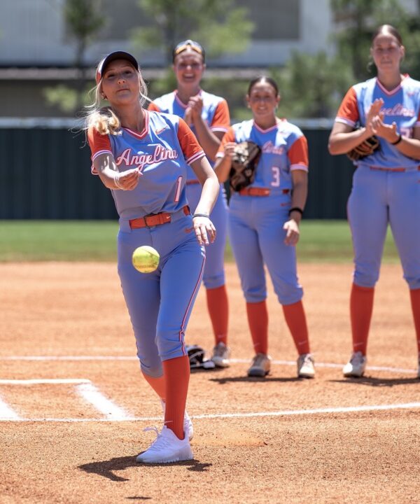 Softball player in blue and orange uniform pitches as three teammates behind her clap and watch on the field.