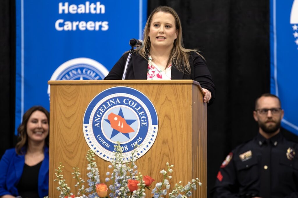 Woman speaks at Angelina College podium, with a woman in blue and an officer beside her. "Health Careers" banners behind.