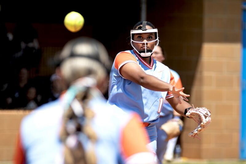 Softball pitcher in blue and orange uniform throws midair ball; focused expression, teammate seen in foreground.