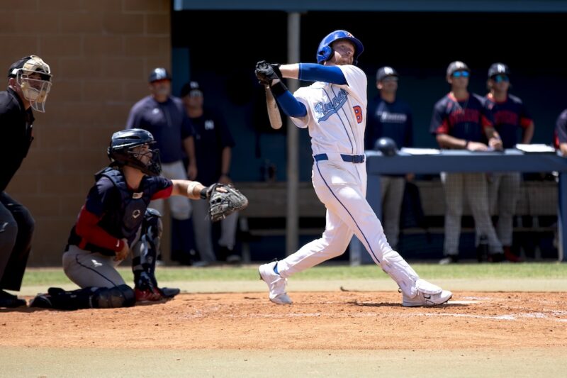 Baseball player in white and blue swings at bat as catcher and umpire watch; teammates stand in dugout behind.