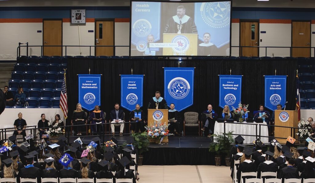 Graduates in caps and gowns sit in a gym facing a stage with a speaker at the podium, faculty, and banners on display.
