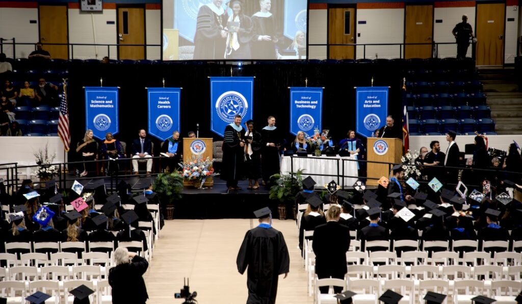 Graduation ceremony in a gym, graduates in caps and gowns, some walking to receive diplomas; banners show academic programs.
