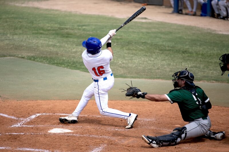 Baseball player in white #16 swings at home plate as catcher in green crouches behind on a sunny field.