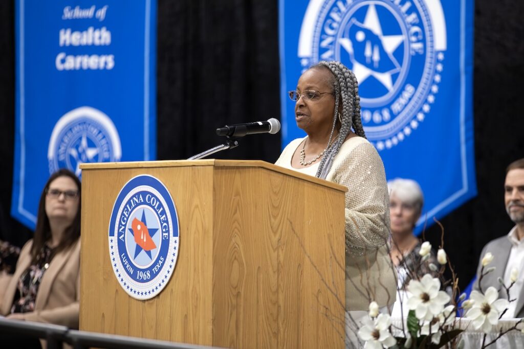 Woman speaks at Angelina College podium, blue banners behind her, white flowers on the podium, audience in view.