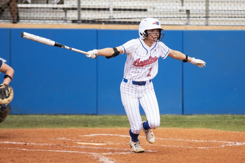 Softball player in white and blue stripes swings bat and runs from home plate; blue fencing and empty bleachers behind.