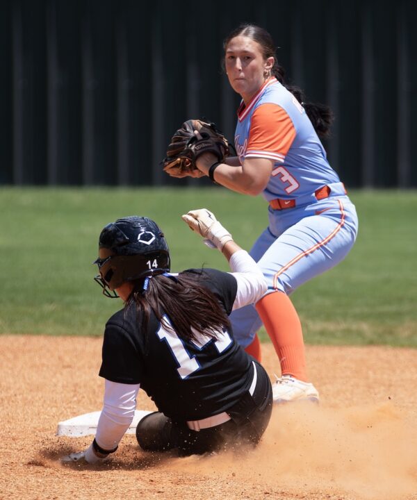 Softball player in blue and orange readies to throw as opposing player slides into base, kicking up dirt on outdoor field.