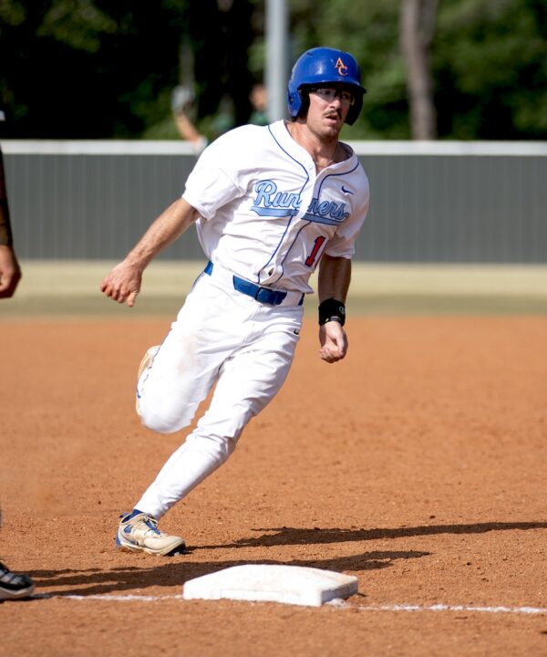 Baseball player in white uniform and blue helmet runs around a base on a dirt field; trees and a fence are in the background.