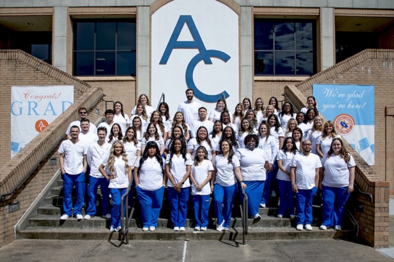 Students in white tops and blue pants celebrate graduation on steps outside a building with an AC sign, banners on both sides.