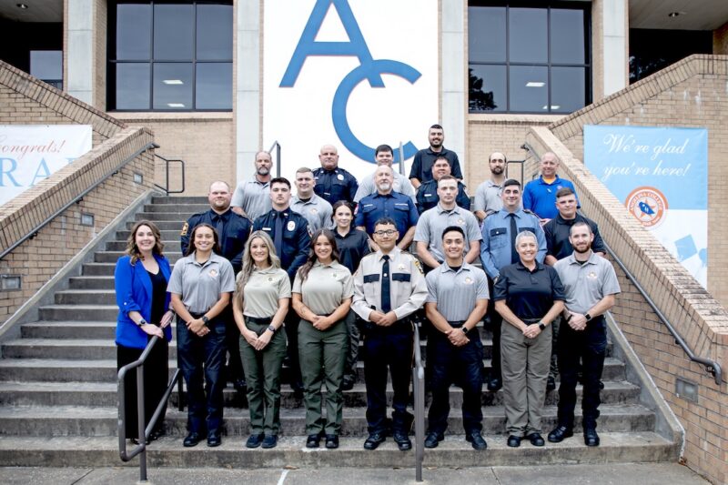 Men and women in uniforms and business attire pose on steps outside a building, with banners on either side of the entrance.