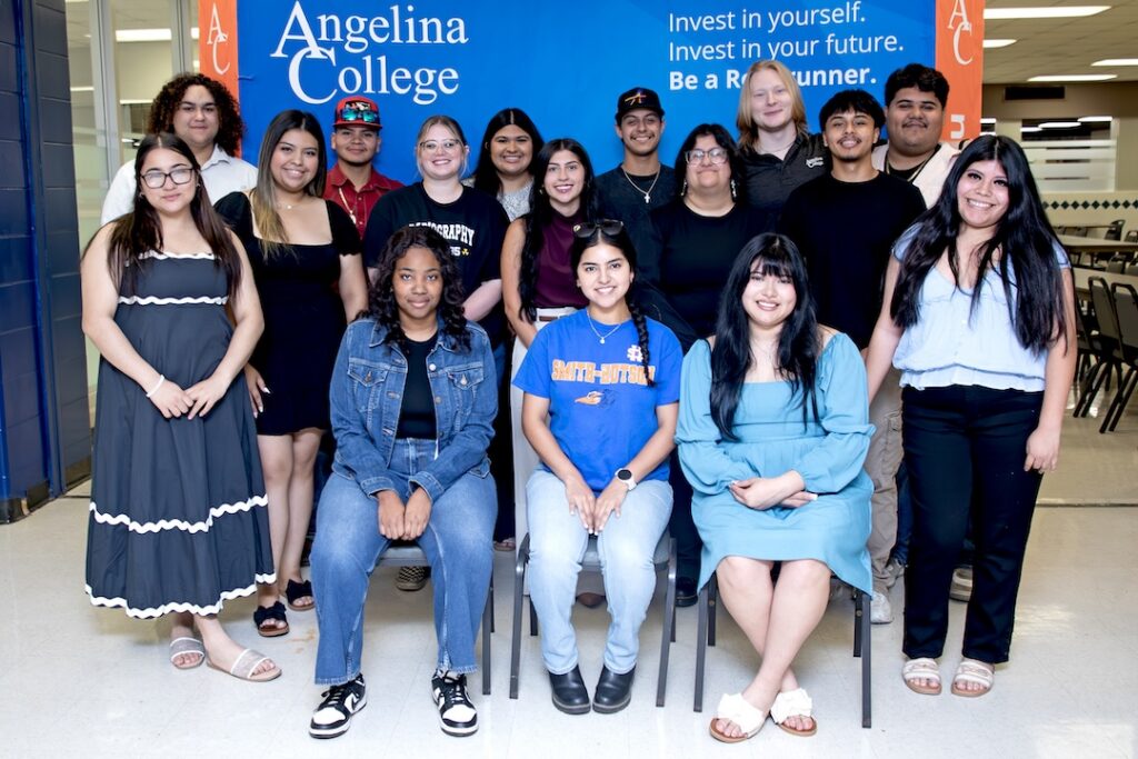 Sixteen smiling students pose in front of an Angelina College banner, some seated and others standing indoors in a common area.