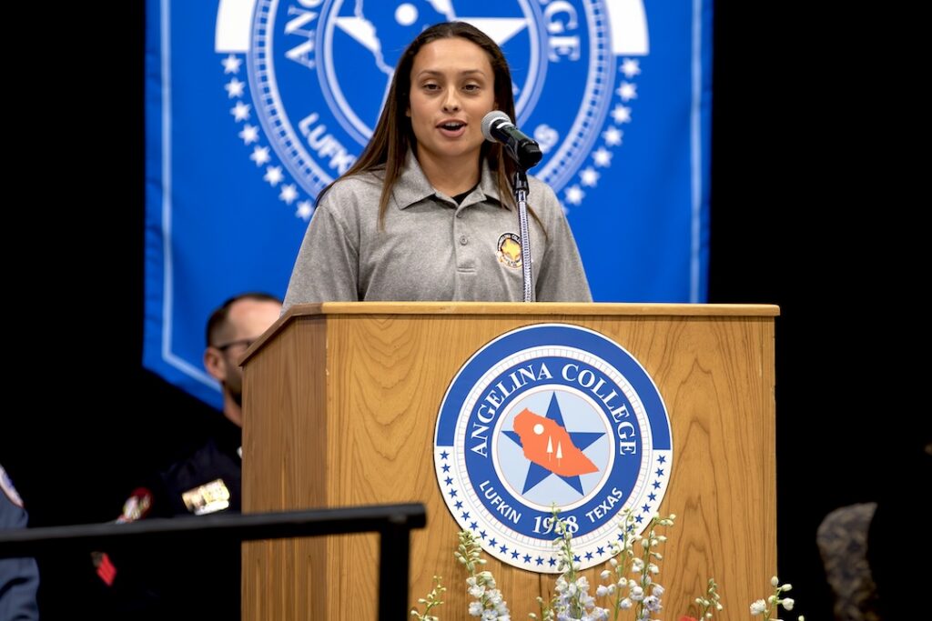 Young woman in gray shirt speaks at Angelina College podium, with flowers, blue banner, and people in uniform behind her.