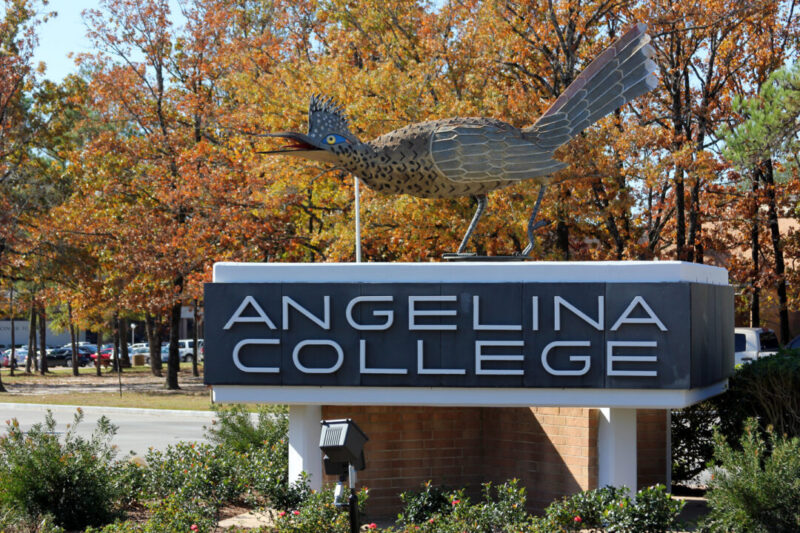 Angelina College sign with bold white letters and a roadrunner sculpture, autumn trees, and parked cars in the background.