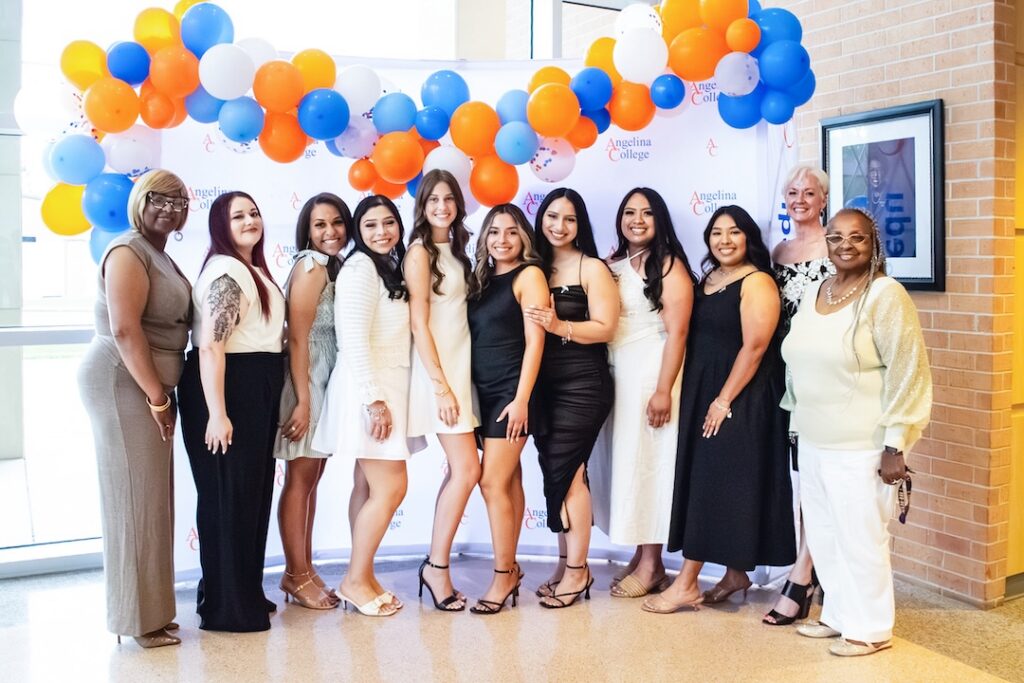 Eleven women stand smiling in front of a white backdrop with blue, orange, and white balloons at a formal Angelina College event.