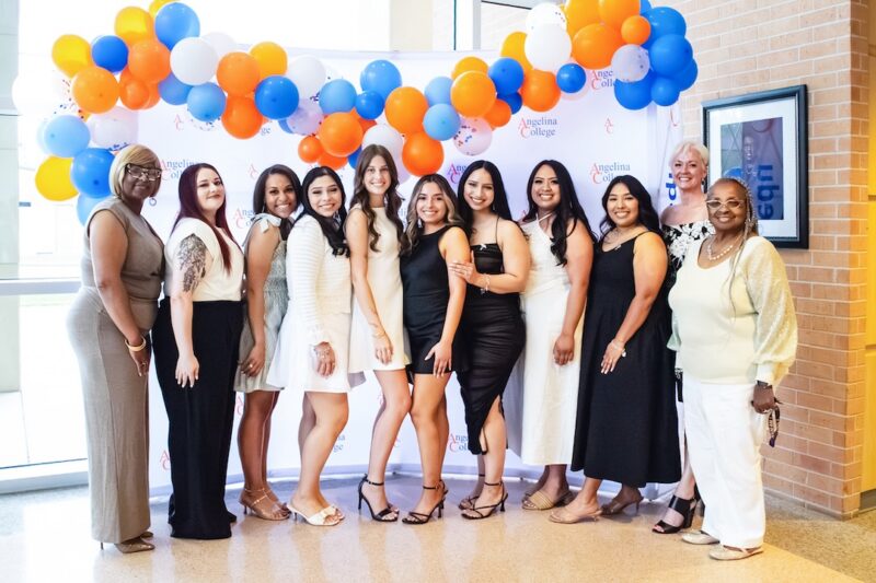 Eleven women stand smiling in front of a white backdrop with blue, orange, and white balloons at a formal Angelina College event.